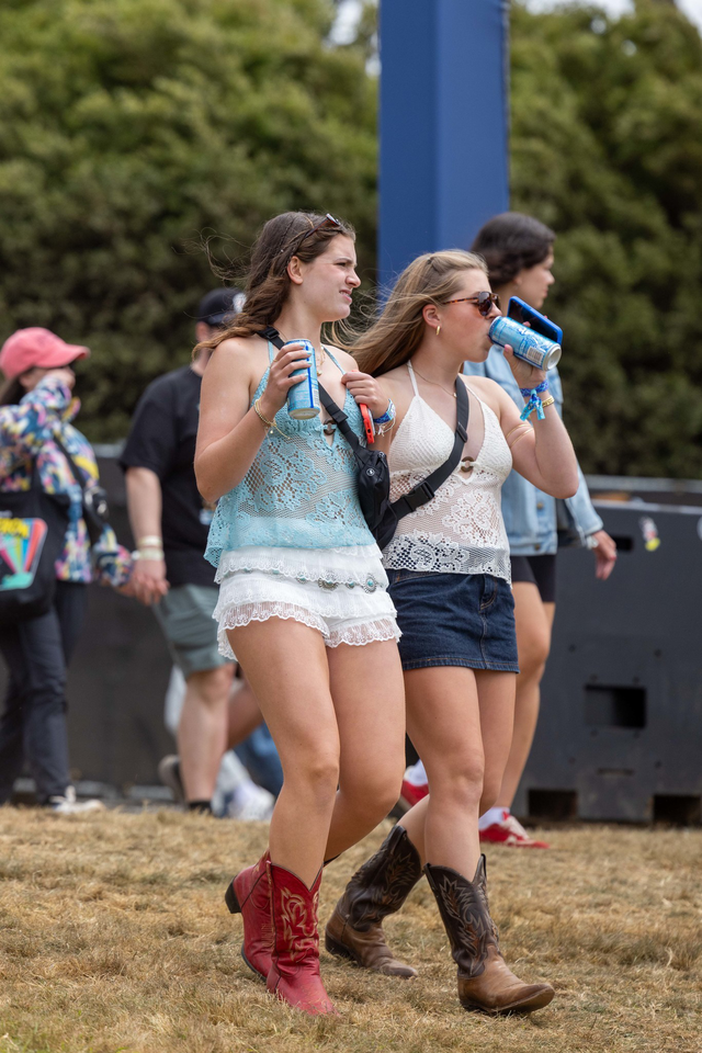 Two young women walk on dry grass, drinking from cans, wearing lace tops, shorts and cowboy boots; people and greenery appear in the background.